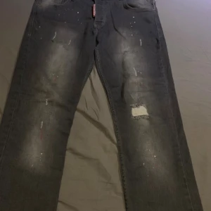 Grå distressed jeans från Dsquared2 - Säljer ett par grå jeans från Dsquared2 med coola slitningar, färgstänk och patchdetaljer. Jeansen har en rak passform och klassisk femficksdesign. Perfekta för dig som gillar en edgy streetstil.