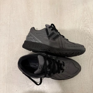 Grå New Balance 1906R sneakers - Säljer ett par grå New Balance 1906R sneakers med svarta detaljer och snörning. Skorna har en sportig vibe med mesh och syntetmaterial, samt robust sula för extra komfort. Perfekta för dig som gillar streetstyle och vill ha något som sticker ut.