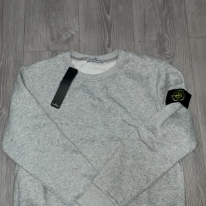 Grå sweatshirt från Stone Island - Snygg grå sweatshirt från Stone Island med klassisk logga på ärmen. Tröjan har rund halsringning och långa ärmar. Perfekt för dig som gillar stilrena och trendiga plagg. Mjuk och skön bomullskvalitet.