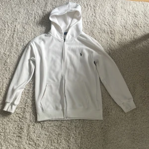 Vit hoodie från Polo Ralph Lauren - Vit hoodie från Polo Ralph Lauren med dragkedja framtill och klassisk huva. Tröjan har två fickor på magen och den ikoniska broderade loggan på bröstet. Tillverkad i mjuk bomull, perfekt för en clean och stilren look.