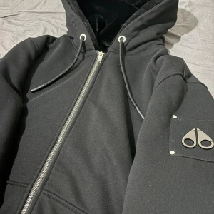 Svart hoodiejacka från Moose Knuckles - Säljer en helt ny och superfräsch Moose Knuckles hoodie-jacka – aldrig använd,perfekt för dig som vill ha en lyxig streetwear-look med riktigt bra kvalité! Passar både L och XL 
