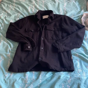 Svart overshirt med knappar - Säljer en svart overshirt med klassisk krage och knäppning framtill. Skjortan har två bröstfickor med lock och knappar, samt långa ärmar med manschett. Perfekt att slänga över en t-shirt för en chill look. Den är i storleken xs men passar även S, jag är 173 cm lång som referens. Går gärna ner i pris lite grann om det låter rimligt