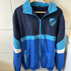Sofialäktaren retrojacka - Retro vindjacka i marinblått och ljusblått med vitt band på ärmarna. Jackan har DIF-logga på bröstet, hel dragkedja framtill och elastiska muddar. Perfekt för dig som vill visa klubbkänsla och ha en sportig stil.