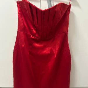 Röd axelbandsklänning från Belle by Oasis - Snygg röd kortklänning från Belle by Oasis med sin  figurnära passform. Klänningen har veckad överdel, dragkedja bak och är tillverkad i glansigt material som ger en lyxig känsla. Perfekt för dig som vill sticka ut med en klassisk och elegant look.