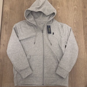 Polo Ralph Lauren Hoodie  - Snygg grå hoodie från Polo Ralph Lauren med dragkedja och huva. Klassisk design med logga broderad på bröstet och praktiska fickor framtill. Tillverkad i mjuk bomullsmix för maximal komfort. Perfekt för en chill och stilren look. Nytt skick storlek S