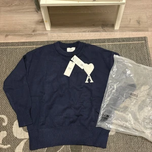 Mörkblå stickad tröja från Ami - Mörkblå stickad tröja från Ami med rund halsringning och ribbade muddar. På bröstet finns ett vitt hjärta med bokstaven A. Tröjan har en klassisk, lite loose passform och är perfekt för lager-på-lager under höst och vinter.