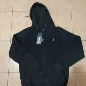 Mörkblå hoodie från Polo Ralph Lauren - Mörkblå hoodie från Polo Ralph Lauren med dragkedja framtill och klassisk vit logga på bröstet. Tröjan har huva med snörning och två fickor framtill. Perfekt för en chill och stilren look.