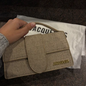 Beige handväska från Jacquemus - Snygg beige handväska från Jacquemus med stilren, rektangulär form och guldfärgad logga. Väskan är tillverkad i linne med detaljer i skinn och har ett praktiskt handtag samt guldfärgade metalldetaljer på sidan. Dustbag och matchande axelrem ingår. Ej äkta. 
