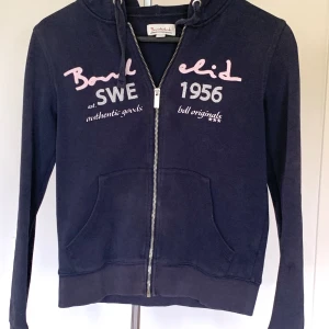 Mörkblå hoodie från Bondelid XS - Snygg mörkblå hoodie från Bondelid med dragkedja och huva. Framsidan har broderade detaljer i ljusrosa och grått med texten 'SWE 1956'. Tröjan har långa ärmar, känguruficka och ribbade muddar. Perfekt för chill dagar och streetstyle.