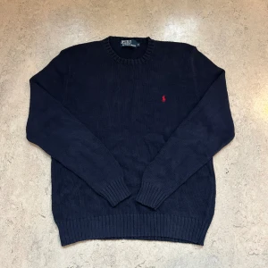 Mörkblå Ralph Lauren stickad tröja  - Säljer en helt ny mörkblå stickad tröja från Polo Ralph Lauren med klassisk rund hals och röd broderad logga på bröstet. Tröjan har ribbade muddar och är långärmad, perfekt för kyligare dagar. Enkel och stilren design som funkar till allt.