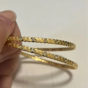 Guldiga armband med glittriga detaljer - Två smala armband i guldton med ingraverade mönster och små glittriga stenar längs hela bandet. Armbanden har en rund form och ger en lyxig känsla med sina skimrande detaljer. Perfekt för dig som gillar att sticka ut med snygga accessoarer.