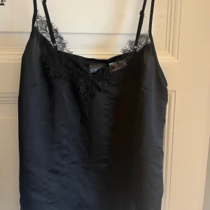 Svart linnetopp med spets från H&M - Säljer en svart linnetopp från H&M i storlek XS. Toppen har tunna axelband och v-ringning med dekorativ spets längs kanten. Materialet är glansigt och känns lyxigt, perfekt för att lyfta vilken outfit som helst.