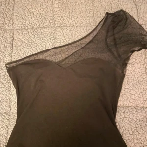 Svart oneshoulder topp med mesh - Snygg svart topp med oneshoulder-design och transparent meshparti upptill. Toppen har en tight passform och är perfekt för dig som vill sticka ut med en unik och trendig look. Den har kort längd och är tillverkad i ett stretchigt material.