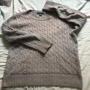 Ralph lauren knit - Kabelstickad grå tröja från Polo Ralph Lauren med rund halsringning och diskret broderad logga på bröstet. Tröjan har långa ärmar, breda ribbade muddar och är tillverkad i mjukt material som passar perfekt för kyligare dagar. Tröjan är S och medium