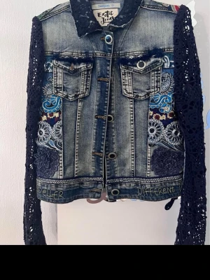 Unik jeansjacka med broderi och spets DESIGUAL  - Säljer en cool jeansjacka från Exotic Jeans med mörkblå virkade spetsärmar och broderade detaljer i blått och vitt. Jackan har knappar framtill, två bröstfickor och textbroderi längst ner. Perfekt för dig som vill sticka ut med din stil!