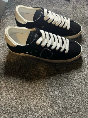 Philippe Model svarta sneakers mocka - Svarta Philippe Model sneakers i mocka med vita detaljer och vita snören. Skorna har en klassisk låg profil, rund tå och vit sula. Märkets logga syns på sidan och hälen är i vitt skinn. Perfekta för dig som gillar stilrena och trendiga sneakers. Storlek 42