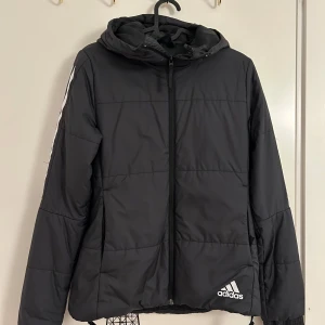 Svart dunjacka från Adidas - Snygg svart dunjacka från Adidas med huva och dragkedja i storlek XS. Jackan har vita detaljer på ärmarna och Adidas-logga på framsidan. Använd ett fåtal gånger och är i mycket bra skick. Perfekt för de flesta väder.