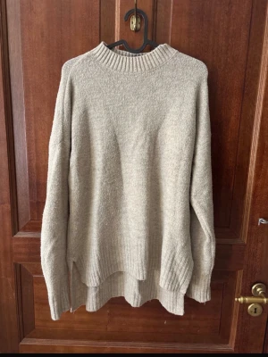 Beige stickad tröja med ribbad kant - Mysig beige stickad tröja med rund hals och ribbade detaljer vid hals, ärmslut och nederkant. Tröjan har en loose passform och är perfekt för kyliga dagar. Enkel och stilren design som passar till det mesta.