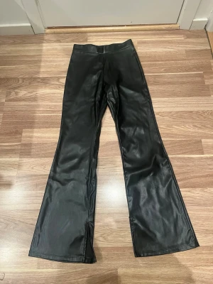 Svarta bootcut byxor i skinnimitation H&M - Snygga svarta bootcut byxor från H&M i skinnimitation. Byxorna har hög midja och slits nertill på benen för en extra cool look. Perfekta för dig som vill sticka ut med en trendig och edgy stil.