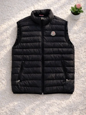 Svart dunväst från Moncler - Säljer en Moncler väst i storlek M. Nypris 3000 kr+.  Västen är i mycket fint skick och sparsamt använd. Alla originaltaggar finns kvar. Det enda att anmärka på är att texten i innerfodret/linningen har gått av, vilket syns tydligt på bilden – inget som påverkar funktion eller användning.  En stilren och varm väst som passar perfekt till vardags eller lager-på-lager.   📏 Storlek: M 💰 Nypris: 3000 kr+ 📦 Skick: Mycket fint (se anmärkning ovan)  Hör gärna av dig vid frågor eller funderingar!