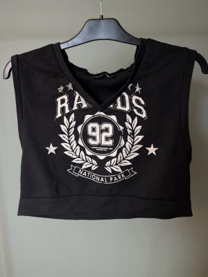 Svart croppad linnetopp FB Sister - Cool svart croppad linnetopp från FB Sister med vitt tryck framtill, texten 'RAIDS 92 NATIONAL PARK' och stjärndetaljer. Toppen har v-ringning och är ärmlös, perfekt till jeans eller shorts för en avslappnad look.