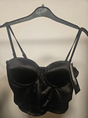 Svart satin korsett från Amisu  - Snygg svart korsett i satin från Amisu med smala axelband och markerade kupor. Toppen har en guldig dragkedja bak och en croppad passform som ger en edgy look. Perfekt att styla med jeans eller kjol för en cool vibe. Lapparna är kvar, den är oanvänd i storlek Small. Går att ta av banden.