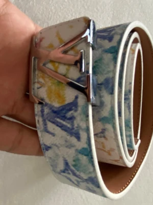 Iv bälte med färgstänk - Iv bälte i vitt skinn med färgstänk i blått, gult och grönt. Snyggt Iv-spänne i silverfärgad metall och klassisk design. Bältet har en unik konstnärlig look och är tillverkat i Spanien. Perfekt för dig som vill sticka ut med accessoarer.