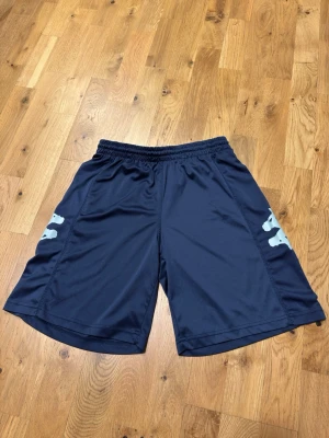 Marinblå träningsshorts med tryck - Säljer ett par marinblå träningsshorts i syntetmaterial med elastisk midja och vita tryck på sidorna. Shortsen har en loose passform och är perfekta för sport eller gym. Materialet är lätt och andas, vilket gör dem sköna att röra sig i.