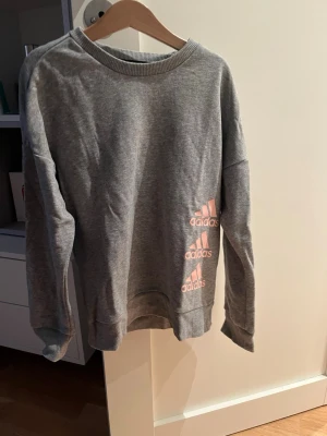 Grå sweatshirt från Adidas - Säljer en grå sweatshirt från Adidas med rosa loggor tryckta på framsidan. Tröjan har rund halsringning, långa ärmar och är tillverkad i mjuk bomullsmix. Perfekt för chill dagar eller till träning.