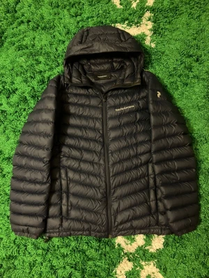 Peak Performance Dunjacka - Säljer en mörkblå dunjacka från Peak Performance med huva och dragkedja. Jackan har ett quiltat mönster och logga på bröstet samt ärmen. Perfekt för kalla dagar och riktigt skön att bära. Passar dig som vill ha en stilren och funktionell jacka. Storlek S. 90% ankdun 10% ankfjäder. Jackan är i väldigt fint skick och har inga täcken på användning. 