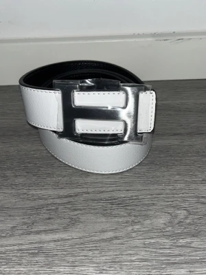 H-bälte  - Snyggt bälte i vitt och svart skinn med ikoniskt,H-spänne i silverfärgad metall. Bältet har stilrena, kontrastsömmar och klassisk design som lyfter, vilken outfit som helst. Strl 110cm
