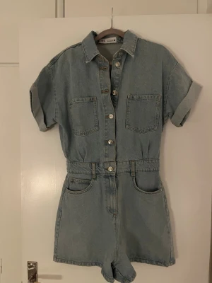 Ljusblå jeansbyxdress från Zara - Säljer en ljusblå jeansbyxdress från Zara i storlek S. Byxdressen har korta ärmar med uppvik, två bröstfickor, knappar framtill och shortsmodell. Perfekt till sommaren och enkel att styla med sneakers eller sandaler.