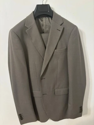 Grå kostym från Riley Stanton - Säljer en stilren grå kostym från Riley Stanton. Kavajen har klassisk passform med två knappar, fyra knappar vid ärmslut och snygga fickdetaljer. Byxorna är raka och har pressveck. Materialet är mjukt och känns lyxigt, perfekt till mer uppklädda tillfällen.