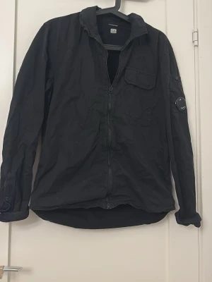 Svart overshirt från C.P. Company - Säljer en svart overshirt från C.P. Company med dragkedja framtill och en ficka på bröstet. Jackan har lång ärm och den ikoniska linsen på ärmen. Perfekt för dig som gillar stilrena och funktionella plagg.
