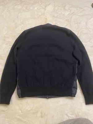 Svart quiltad pufferjacka från Moncler - Säljer en svart pufferjacka från Moncler med quiltad front och rygg i blankt material och stickade ärmar. Jackan har dragkedja framtill, två sidofickor med dragkedja och Moncler-logga på ärmen. Perfekt för dig som vill ha en stilren och modern look.