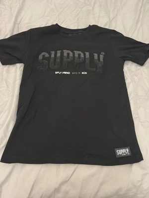 Svart SUPPLY t-shirt med tryck - Snygg svart t-shirt från SUPPLY med stort texttryck på bröstet och en liten patch nertill. Klassisk passform och rund hals. Perfekt till jeans eller shorts för en clean streetwear-look.