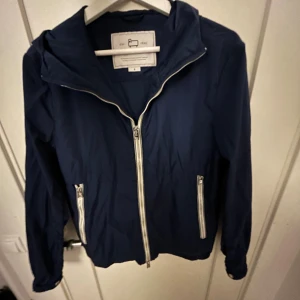 Blå vindjacka från Woolrich - Säljer en blå vindjacka från Woolrich med huva och vita dragkedjor. Jackan har två fickor med dragkedja och är tillverkad i lätt polyester. Perfekt för blåsiga dagar och enkel att matcha med olika outfits. Jackan är köpt från deras affär i Italien. Jackan är använd 2-3 gånger.En sällsynt modell och kostar ca 4000-5000 nypris. Priset är inte hugget i sten. Bara att höra av sig vid minsta fundering.