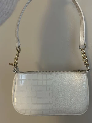 Vit axelväska med kedjedetaljer - Snygg vit axelväska i croco-mönstrat syntetmaterial med guldfärgade kedjedetaljer på axelremmen. Väskan har dragkedja upptill och en stilren, kompakt form som passar perfekt till alla tillfällen.🤍