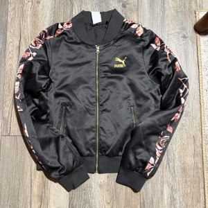 Svart bomberjacka Puma Kenza S - Svart bomberjacka från Puma Kenza med blommigt mönster i rosa och rött längs ärmarna. Jackan har guldfärgad dragkedja, ribbade muddar och två fickor med dragkedja. Materialet är glansigt och känns lyxigt, perfekt för att lyfta din outfit.