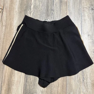 Svarta shorts med vit rand, S från Zara - Svarta shorts med bred midja och en vit rand längs sidan. Shortsen har en mjuk och stretchig känsla, perfekt för en avslappnad och sportig look. Passformen är normal.