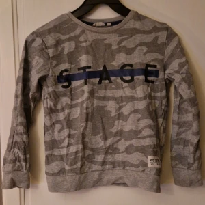 Grå camo tröja från KappAhl - Säljer en grå långärmad tröja från KappAhl med camouflagemönster och texten 'STAGE' över bröstet i svart och blått. Tröjan har ribbade muddar och är gjord i mjuk bomull. Hund finns i hemmet. Kolla in mina andra annonser för samfrakt! 