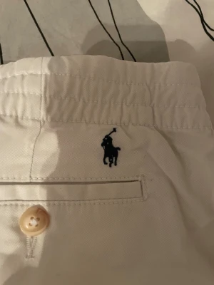 Vita chinos från Polo Ralph Lauren - Snygga vita chinos från Polo Ralph Lauren med elastisk midja och klassisk broderad logga vid bakfickan. Byxorna har raka ben, knappstängning och är gjorda i mjuk bomull. Perfekta för en clean och stilren look.