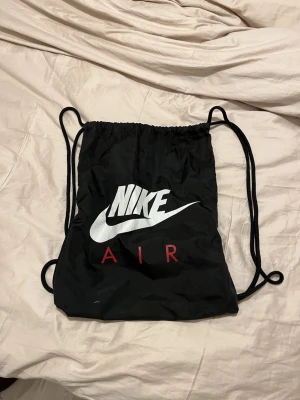 Svart Nike Air gympåse - Säljer en svart gympåse från Nike Air med stor vit logga och röd text. Väskan har snörning upptill och svarta axelband. Perfekt för att bära träningskläder eller skor. Tillverkad i lätt syntetmaterial.
