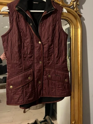 Vinröd quiltad Barbour dunväst - Säljer en vinröd quiltad dunväst från Barbour med guldfärgade tryckknappar och två stora fickor framtill. Västen har dragkedja och hög krage med svart fleecefoder på insidan. Perfekt att slänga över en hoodie eller tröja för extra värme.
