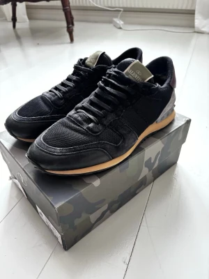 Valentino Rockrunners - Svarta Valentino Rockrunners sneakers med mesh och mockadetaljer, beige sula och vinröda partier på hälen. Klassisk snörning och cool design med diskret logga på plösen. Perfekta för dig som vill ha en lyxig och stilren sneaker med unika detaljer. Skick 9/10. Originallåda medföljer