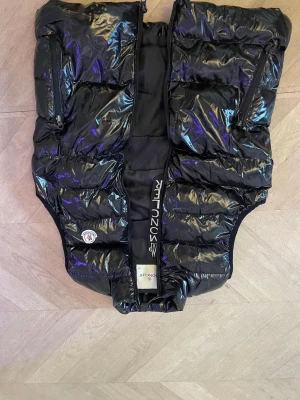Svart glansig dunväst från Moncler - Säljer en svart glansig dunväst från Moncler med klassisk logga på bröstet. Västen har två dragkedjefickor framtill och quiltad design. Perfekt för lager-på-lager och ger en cool vibe till din outfit.