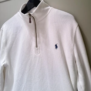Vit half zip tröja från Polo Ralph Lauren - Vit långärmad tröja från Polo Ralph Lauren med half zip och klassisk ståkrage. Tröjan har det ikoniska blå broderade Polo-logot på bröstet och är tillverkad i mjuk bomull. Perfekt för en clean och stilren look.