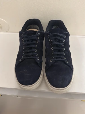 Mörkblå mockasneakers från Sandö - Snygga mörkblå sneakers från Sandö i mocka med vita, tjocka gummisulor. Skorna har klassisk snörning med metallhål och en stilren, låg profil som passar till det mesta. Perfekta för dig som gillar en clean och enkel look.