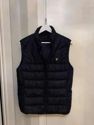 Blå dunväst från Lyle & Scott - Säljer en svart dunväst från Lyle & Scott med gul logga på bröstet. Västen har hög krage, quiltad design och dragkedja framtill. Perfekt att slänga över en hoodie eller tröja för extra värme. Klassisk och stilren look. Inga slitningar eller tecken på användning. Använd bara ett fåtal gånger. 