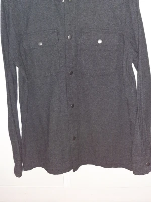 Mörkgrå overshirt från Kappahl - Snygg mörkgrå overshirt från Kappahl i 100% bomull. Jackan har två stora bröstfickor med knappar och tryckknappar längs hela framsidan. Perfekt lager-på-lager-plagg med lång ärm och stilren look. längd- 74, bred-55 cm